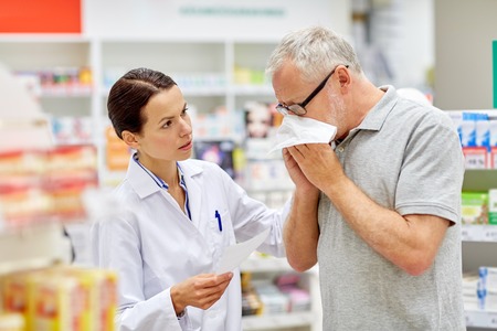 medicine, pharmaceutics, health care and people concept - pharmacist and sick senior man with flu blowing nose at pharmacy