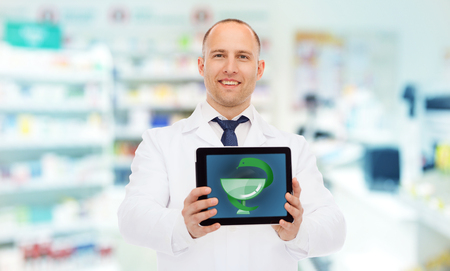 medicine, pharmacy, people, health care and pharmacology concept - smiling male doctor showing tablet pc computer with medical symbol of snake on screen and cup over drugstore backgroundの写真素材
