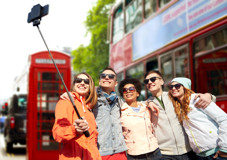 tourism, travel, people, leisure and technology concept - group of smiling teenage friends taking selfie with smartphone and monopod over london city street backgroundの写真素材