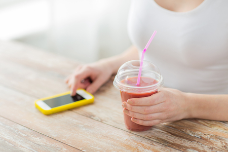 healthy eating, diet, technology and people concept - close up of woman with smartphone and cup of smoothie sitting at tableの写真素材