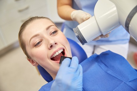 people, medicine, stomatology, technology and health care concept - close up of dentist putting intraoral shield to female patient mouth and assistant directing x-ray machine at dental clinicの写真素材