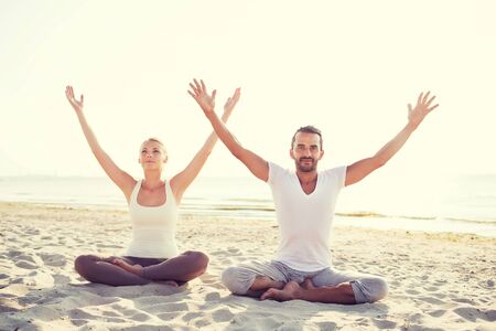 fitness, sport, friendship and lifestyle concept - smiling couple making yoga exercises sitting on sand outdoorsの写真素材