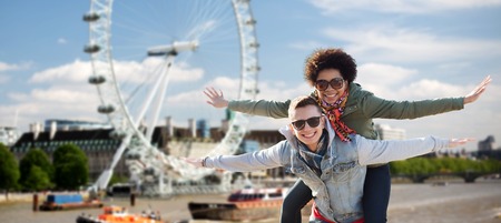 friendship, leisure, international, freedom and people concept - happy teenage couple in shades having fun over london ferry wheel backgroundの写真素材