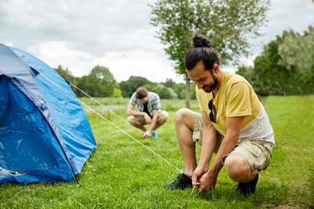camping, travel, tourism, hike and people concept - smiling friends setting up tent outdoorsの写真素材