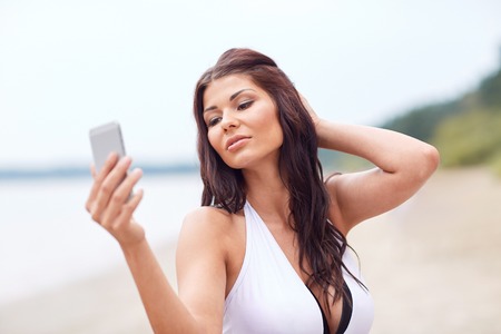 summer, travel, technology and people concept - sexy young woman taking selfie with smartphone on beachの写真素材