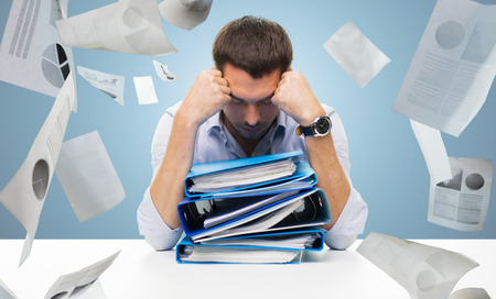 business, people, deadline, stress and paperwork concept - sad businessman with stack of folders and falling papers over blue backgroundの写真素材