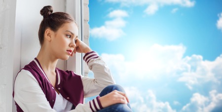 people, emotion and teens concept - sad unhappy pretty teenage girl sitting on windowsill over blue sky and clouds backgroundの写真素材