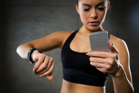 sport, fitness, technology and people concept - close up of young woman with heart-rate or smart watch and smartphone in gymの写真素材