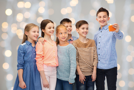 childhood, friendship, technology and people concept - happy children talking selfie by smartphone over holidays lights backgroundの写真素材