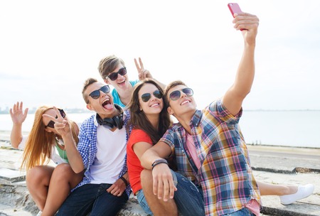 friendship, leisure, summer, technology and people concept - group of happy friends with smartphone...