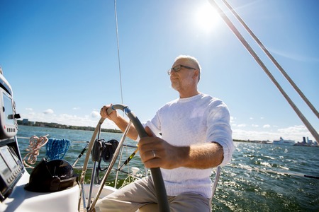 sailing, age, tourism, travel and people concept - happy senior man in captain hat on steering wheel and navigating sail boat or yacht floating in seaの写真素材