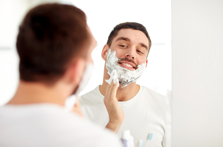 beauty, hygiene, shaving, grooming and people concept - smiling young man looking to mirror and applying shaving foam to face at home bathroomの写真素材
