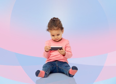 childhood, technology and people concept - smiling little african american baby girl playing with smartphone and sitting on floor over pink violet backgroundの写真素材