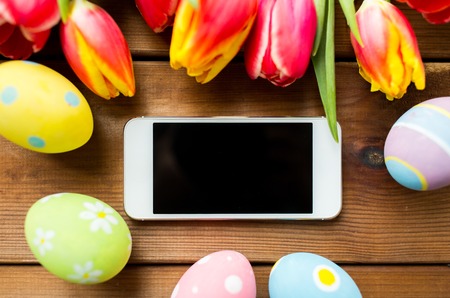 easter, holidays, tradition and object concept - close up of colored easter eggs, tulip flowers and smartphone with blank screen on wooden surfaceの写真素材