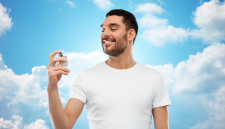 perfumery, beauty and people concept - happy smiling young man with male perfume over blue sky and clouds backgroundの写真素材
