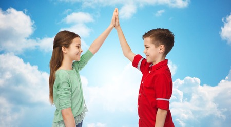 childhood, friendship, gesture and people concept - happy smiling boy and girl making high five over blue sky and clouds backgroundの写真素材
