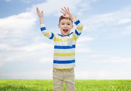 childhood, fashion, joy and people concept - happy little boy waving hands over blue sky and green...