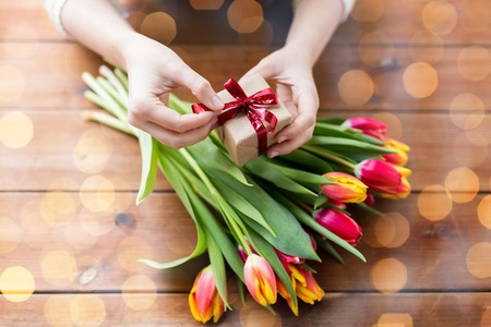 people, holidays and greeting concept - close up of woman holding gift box and tulip flowers over...