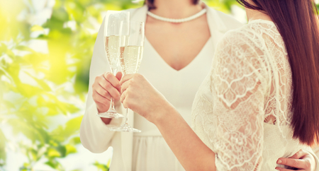 people, homosexuality, same-sex marriage, celebration and love concept - close up of happy married lesbian couple holding and clinking champagne glasses over green tree leavers backgroundの写真素材