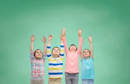 childhood, school, education, gesture and people concept - happy smiling children raising hands and celebrating victory over green school chalk board backgroundの写真素材