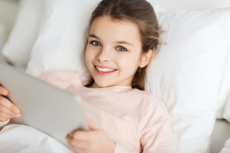 people, children, rest and technology concept - happy smiling girl lying awake with tablet pc computer in bed at homeの写真素材
