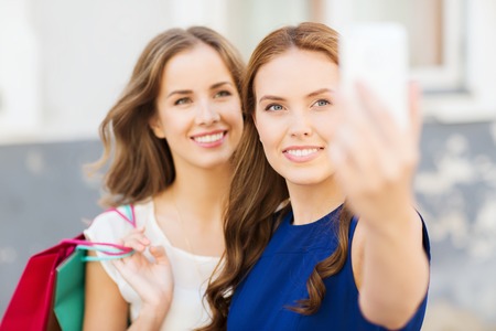 sale, consumerism, technology and people concept - happy young women with shopping bags and smartphone taking selfie on city streetの写真素材