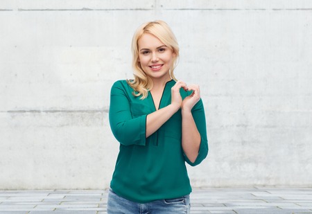 love, valentines day, gesture, plus size and people concept - smiling young woman in shirt and jeans showing heart shape hand sign over gray concrete wall backgroundの写真素材