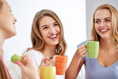 friendship, people, pajama party and communication concept - happy friends or teenage girls drinking, chatting tea and eating sweets at homeの写真素材