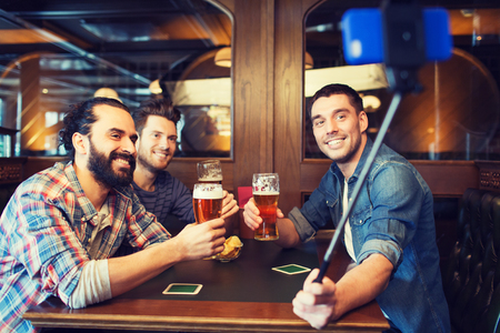 people, men, leisure, friendship and technology concept - happy male friends drinking beer and taking picture with smartphone selfie stick at bar or pubの写真素材