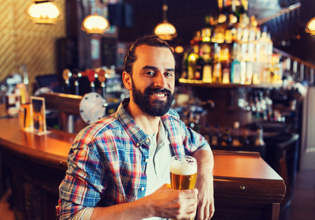 people, drinks, alcohol and leisure concept - happy young man drinking beer at bar or pubの写真素材