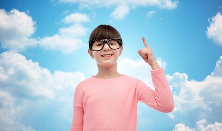 childhood, school, education, vision and people concept - happy little girl in eyeglasses pointing finger up over blue sky and clouds backgroundの写真素材