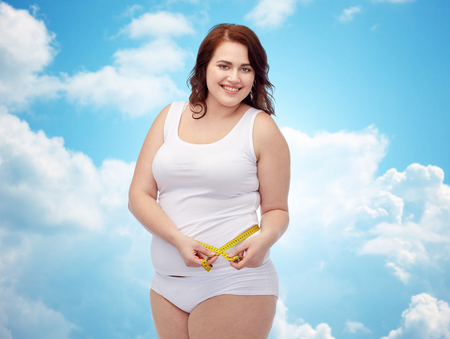 weight loss, diet, slimming, size and people concept - happy young plus size woman in underwear measuring tape over blue sky and clouds backgroundの写真素材
