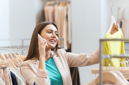 sale, consumerism, fashion, communication and people concept - happy young woman with shopping bags choosing clothes and calling on smartphone in mall or clothing storeの写真素材