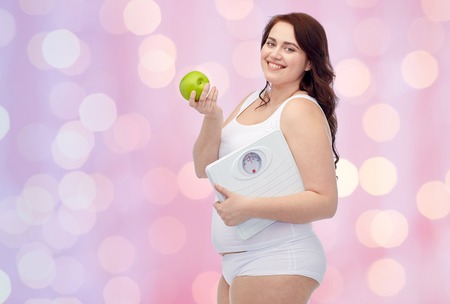 weight loss, diet, slimming, healthy eating and people concept - happy young plus size woman in underwear holding scales and green apple over pink holidays lights backgroundの写真素材