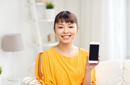 people, technology, communication and leisure concept - happy young asian woman sitting on sofa and showing smartphone blank screen at homeの写真素材
