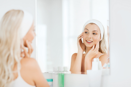 beauty, skin care and people concept - smiling young woman in hairband touching her face and looking to mirror at home bathroomの写真素材