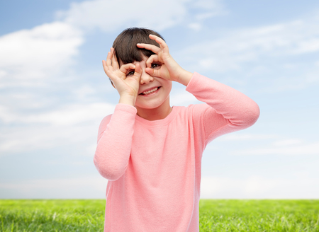 childhood, fun, gesture and people concept - happy little girl making faces over blue sky and grass backgroundの写真素材