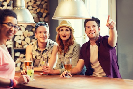 people, leisure, friendship and communication concept - group of happy smiling friends drinking beer and cocktails at bar or pub and pointing finger to somethingの写真素材