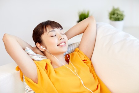 people, technology, relax and leisure concept - happy asian young woman lying on sofa with earphones listening to music at homeの写真素材