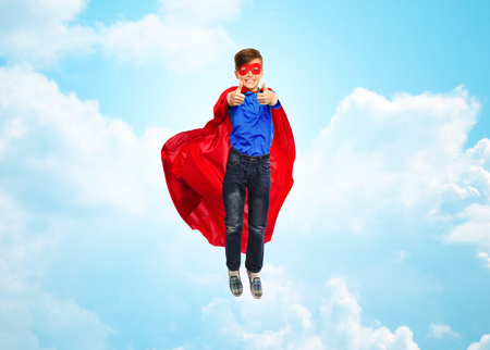 happiness, freedom, childhood, movement and people concept - boy in red super hero cape and mask flying in air and showing thumbs up over blue sky and clouds backgroundの写真素材