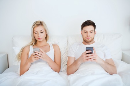 people, technology, internet and communication concept - couple with smartphones in bedの写真素材