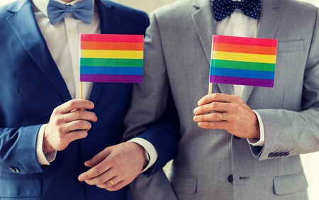 people, homosexuality, same-sex marriage and love concept - close up of happy male gay couple in suits and bow-ties with wedding rings holding rainbow flagsの写真素材