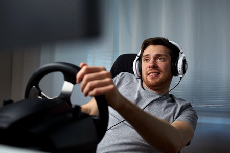 technology, gaming, entertainment and people concept - happy smiling young man in headphones with pc computer playing car racing video game at home and steering wheelの写真素材