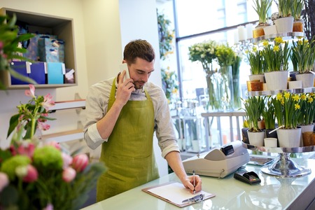people, sale, retail, business and floristry concept - happy smiling florist man calling on smartphone and making notes to clipboard at flower shop counterの写真素材