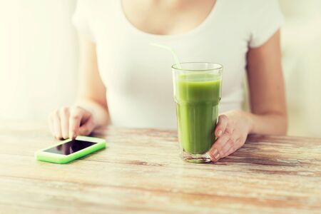 healthy eating, diet, detox, technology and people concept - close up of woman with smartphone green juice sitting at wooden tableの写真素材