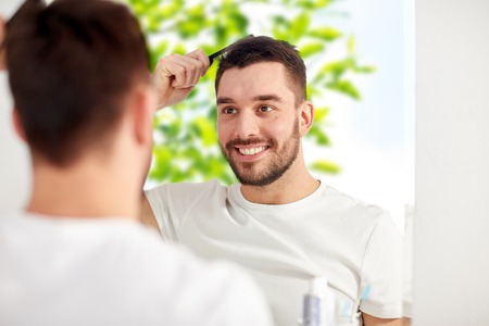 beauty, grooming and people concept - smiling young man looking to mirror and brushing hair with comb at home bathroom over green natural backgroundの写真素材