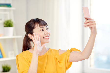 people, technology and leisure concept - happy young asian woman taking selfie on smartphone at home and waving handの写真素材