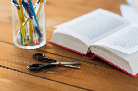 education, school and object concept - close up of stand or glass with writing tools and book with scissors on wooden tableの写真素材