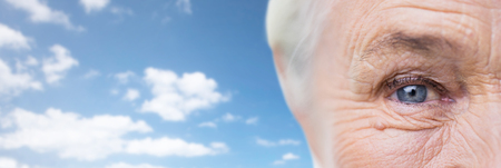 age, vision, eyesight and old people concept - close up of senior woman face and eye over blue sky and clouds backgroundの写真素材