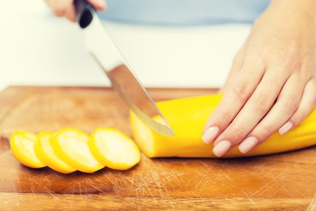 healthy eating, cooking, vegetarian food, vegetable and people concept - close up of woman hands chopping squash with knife on wooden cutting boardの写真素材
